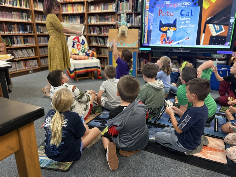 First grade students sit on the carpet and listen to the librarian read a book.
