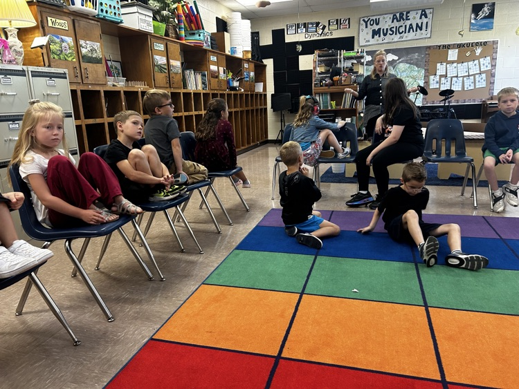     Students listen to their teacher while seated around the music classroom.