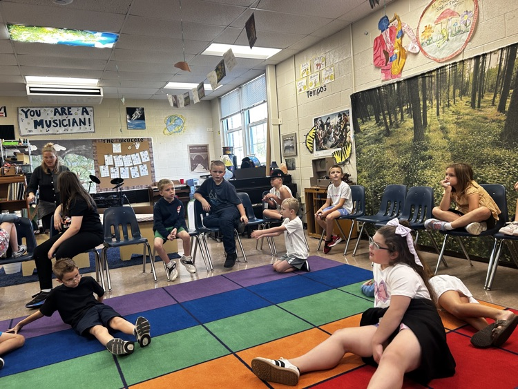 Students sit in a circle on chairs and the colorful rug during a music class activity.