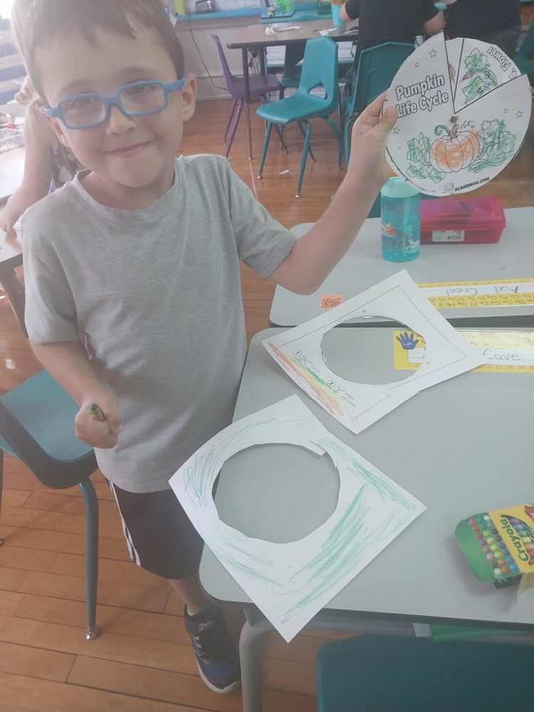 A student in blue glasses smiles and holds up a colored “Pumpkin Life Cycle” wheel in the classroom.