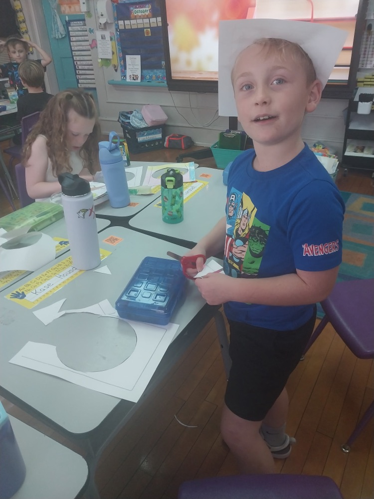 Students work on paper projects at a classroom table. A boy in a blue Avengers shirt cuts paper while a classmate works beside him.