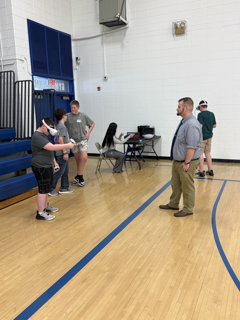 A student wearing a virtual reality headset uses handheld controllers in the gym while other students participate in nearby VR activities.