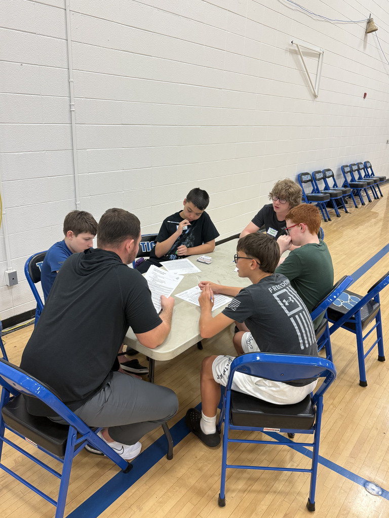 A group of students sits around a table in the gym working through printed activities with an adult facilitator during SPARK 360 Family Transition Night.