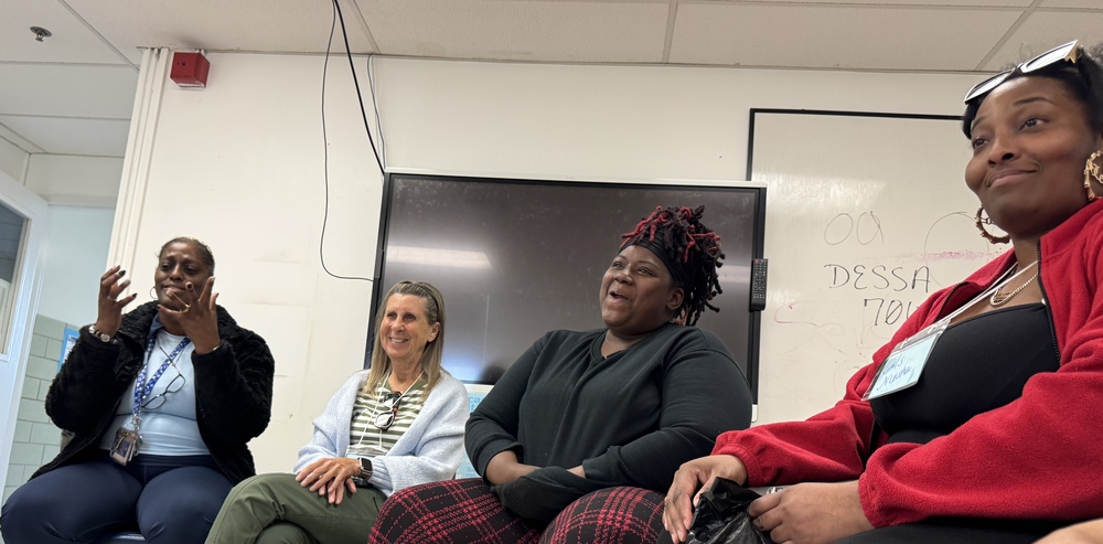 Four women seated in a classroom. One with a watch, another with a black jacket, and two with plaid pants.