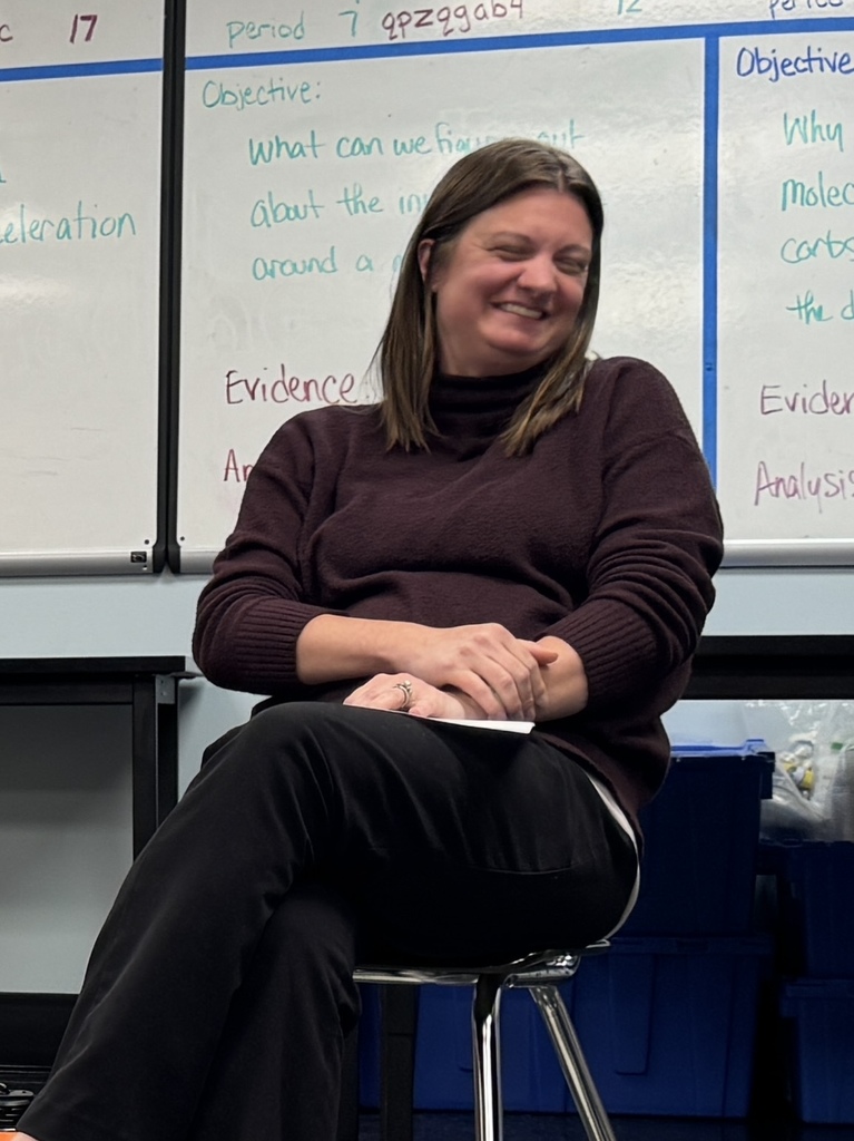 A woman sits in a classroom, smiling, with a whiteboard in the background displaying text and diagrams.