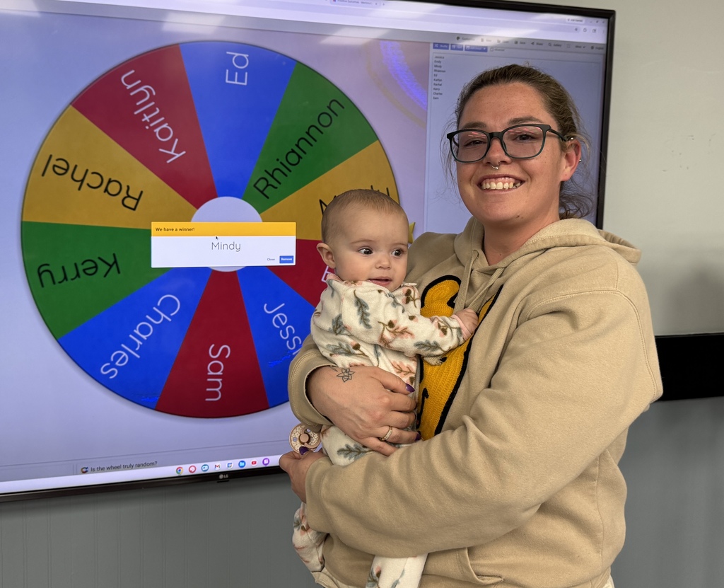 A woman wearing glasses holds a baby in front of a large screen displaying a colorful wheel.