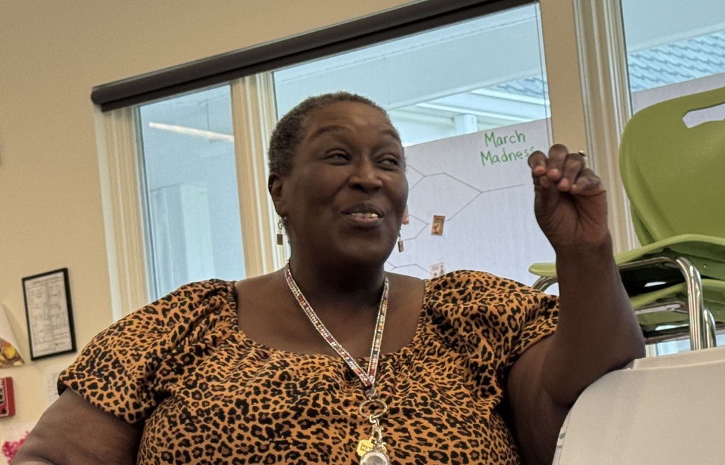 Woman sitting in a classroom, wearing a leopard print blouse and a necklace. Window behind her.
