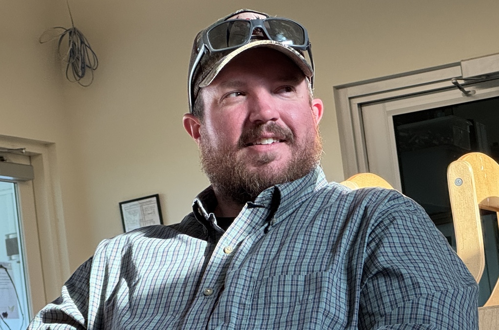 A man with a beard and sunglasses on his cap smiles at the camera in a room.