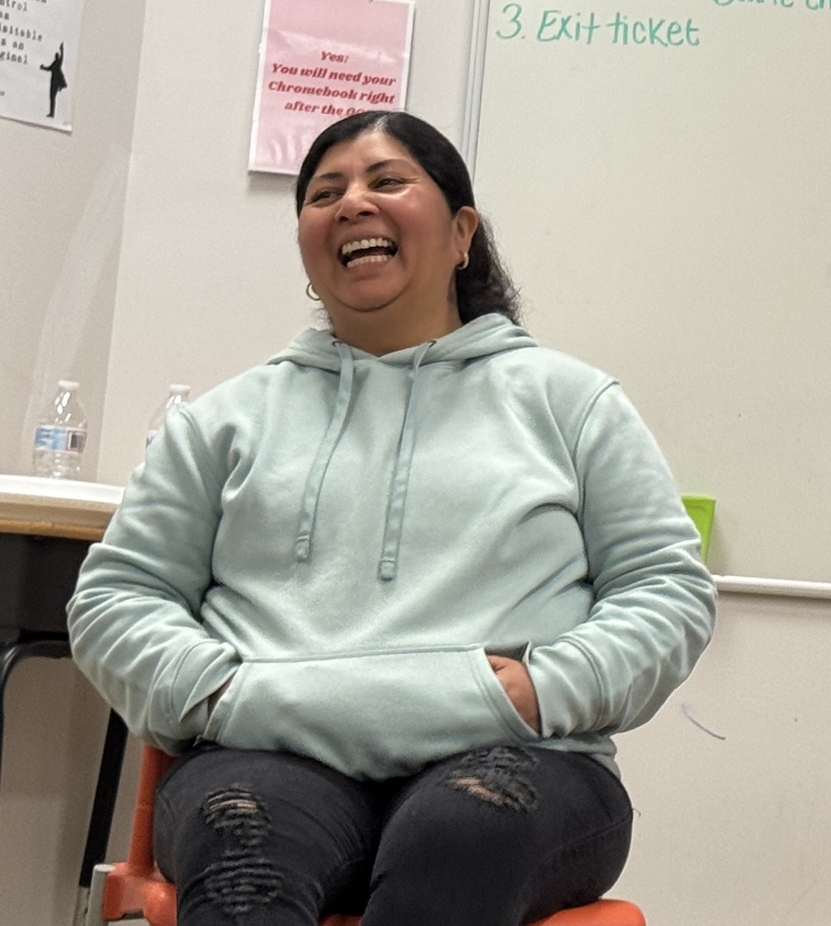 Person in green hoodie, smiling and sitting on a chair, with a whiteboard and classroom setting in the background.