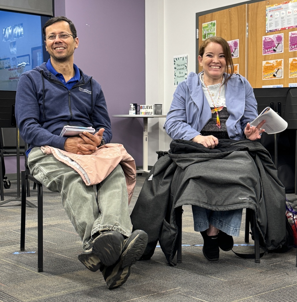 Two people seated in chairs, smiling and holding books, with a bulletin board behind them.