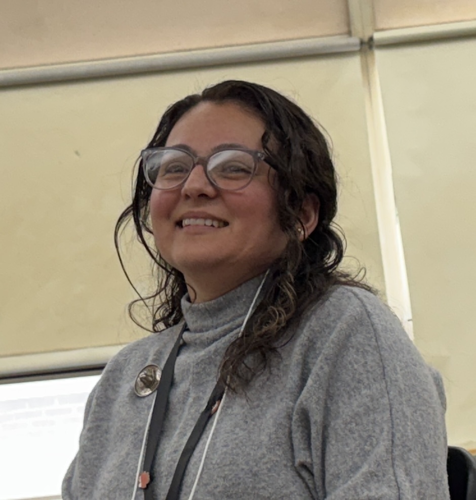 A woman with short dark hair, wearing glasses and a gray top, smiles for the camera. She has a lanyard around her neck.