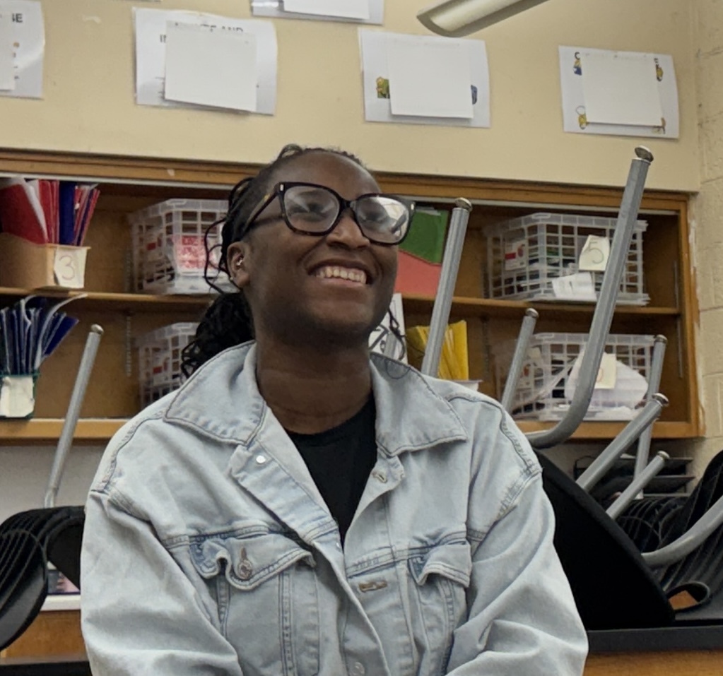 A person in a white jacket with a black shirt smiles in a classroom. Behind them, shelves hold baskets, folders, and other items.