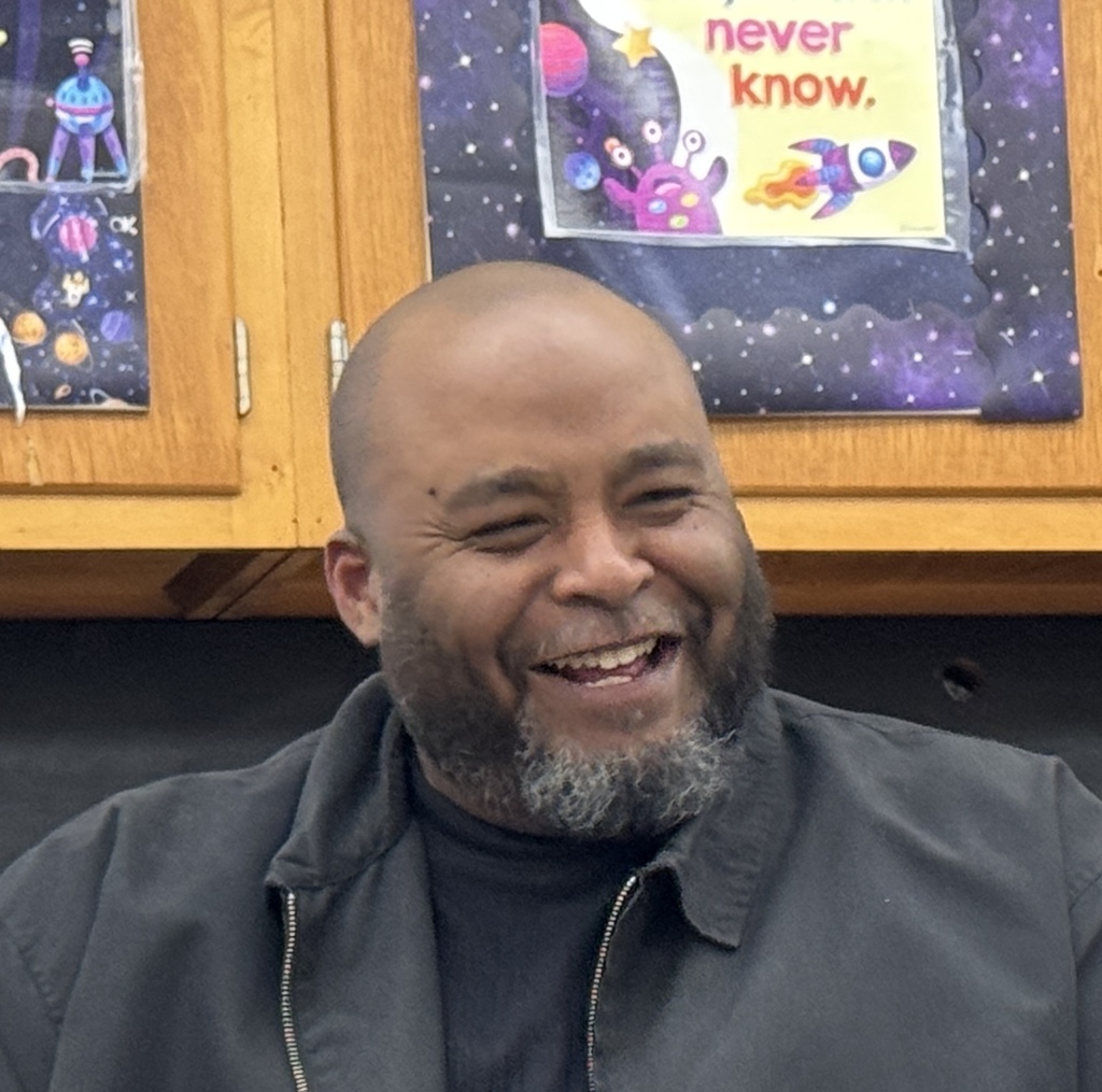 Man smiling with a gray beard and mustache. Behind him are two wooden cabinets with colorful space posters.
