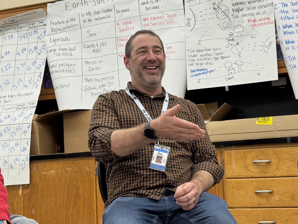 A man sits in a chair, gesturing while smiling, with papers on a wall behind. He wears a brown shirt, jeans, and a watch.