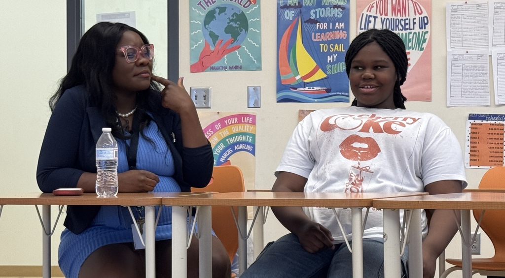 Two individuals seated at a table with a water bottle, one wearing glasses, and a classroom backdrop with posters.