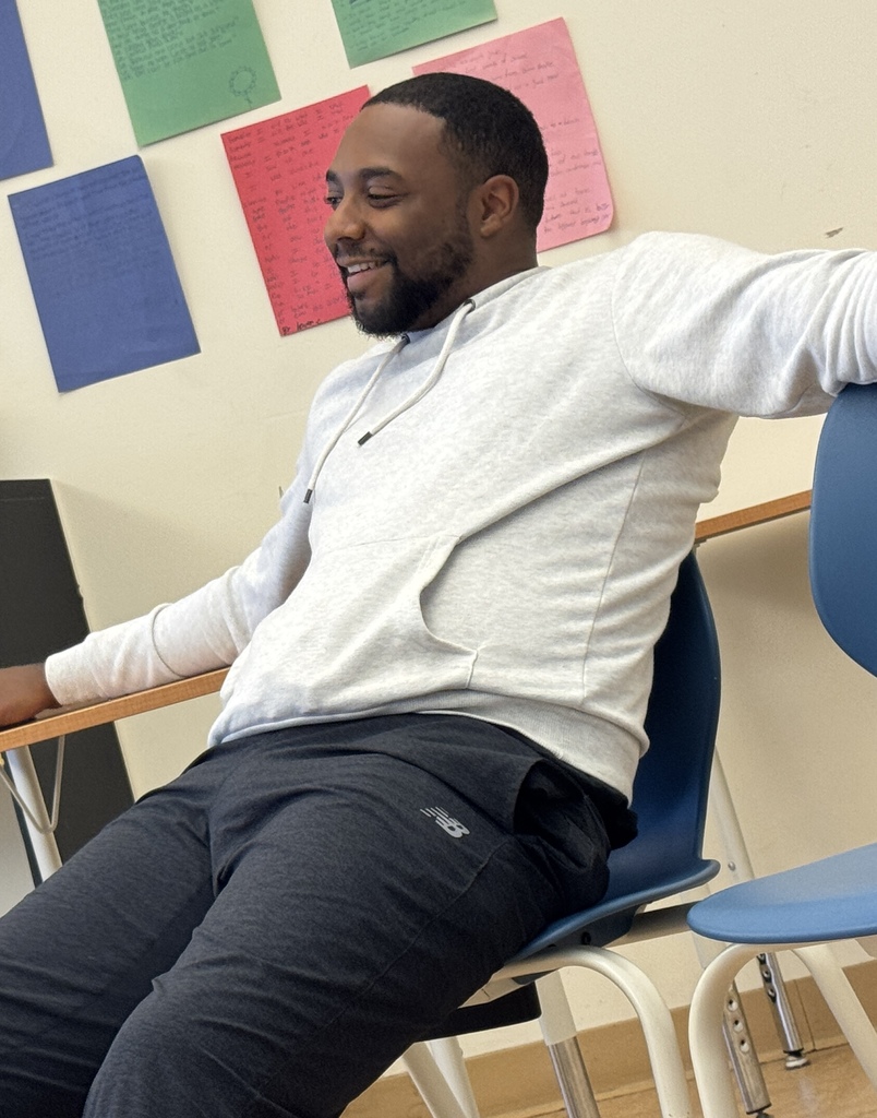 A person in a light-colored hoodie and dark pants sits in a chair, smiling, with colorful papers on the wall behind.
