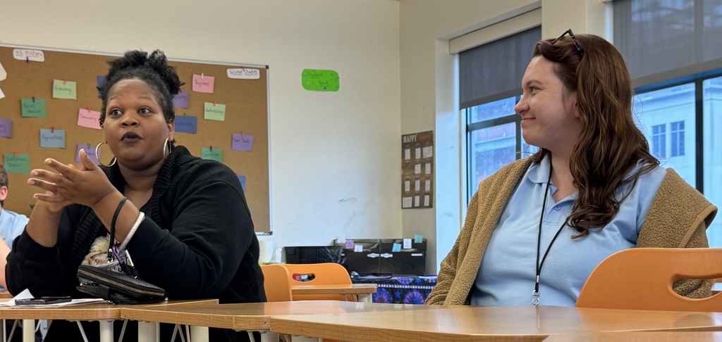 Two women sit at a table in a classroom. One speaks with raised hands. A bulletin board is behind them.