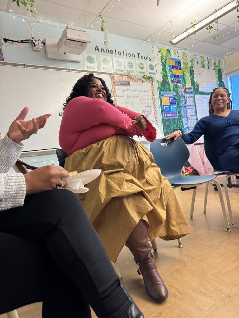 Three women in a classroom, one wearing a yellow skirt and a pink top, sit in chairs. Another person sits on the left, with their hand raised. A whiteboard is behind them.