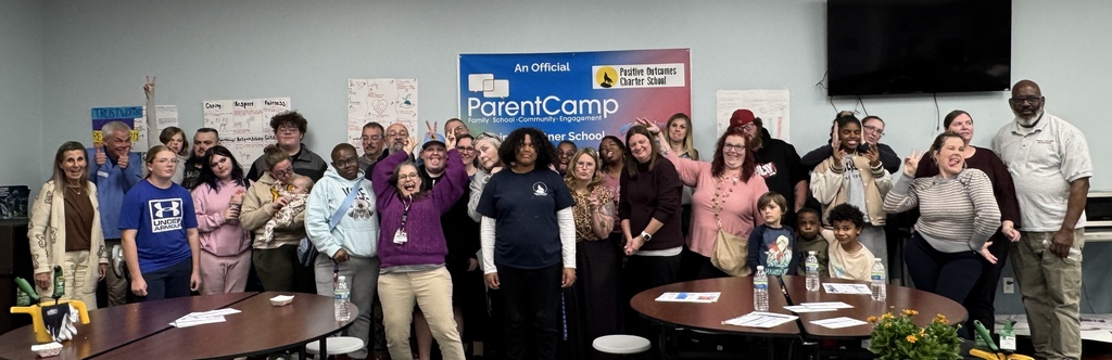 A large group of people gathered in a room, smiling, some standing, some sitting, with tables and a blue banner.