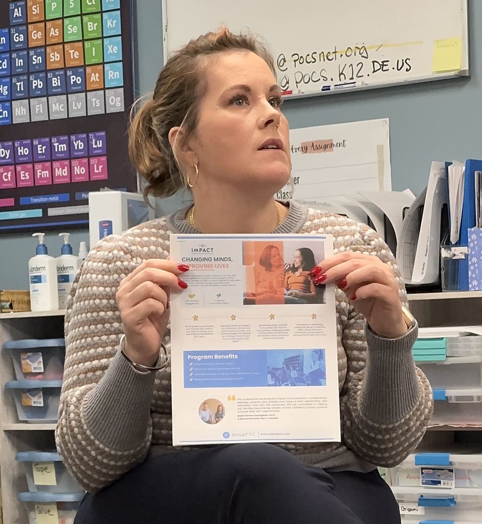 A woman holds a paper in a classroom with a periodic table, whiteboard, and storage boxes.