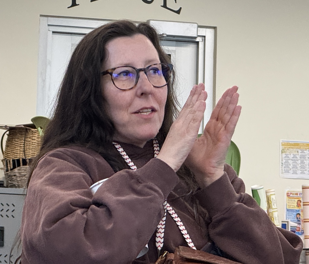 Woman with glasses clapping in a room with beige walls, a door, and items on a shelf.