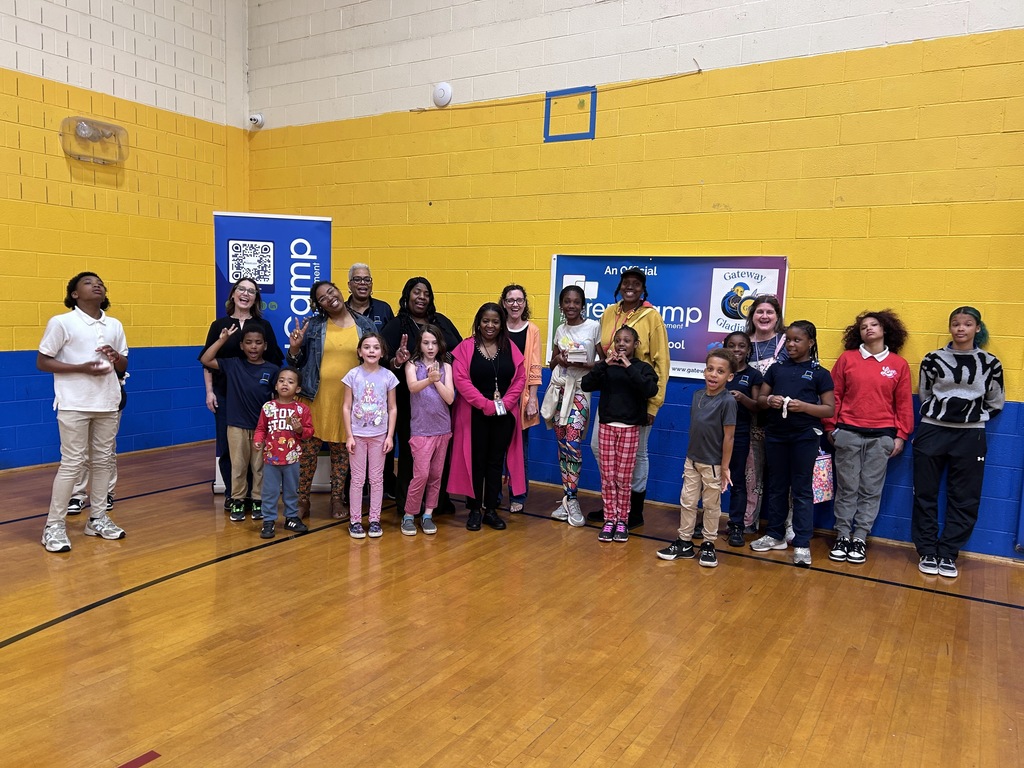 A group of adults and children stand together in a gym with yellow and blue walls.