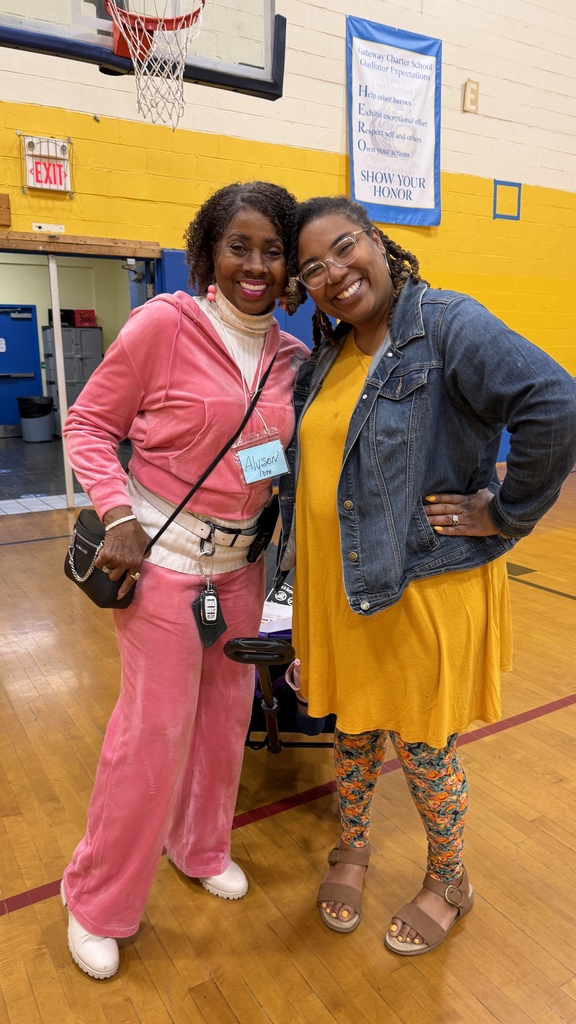 Two women in a gym, one with a pink outfit and the other in yellow, stand smiling. The background includes a basketball hoop and a yellow wall with a banner.