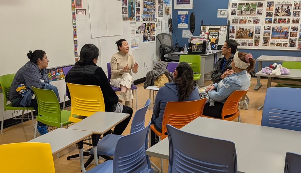 A group of people sit in chairs around tables in a classroom. Some wear winter clothes.