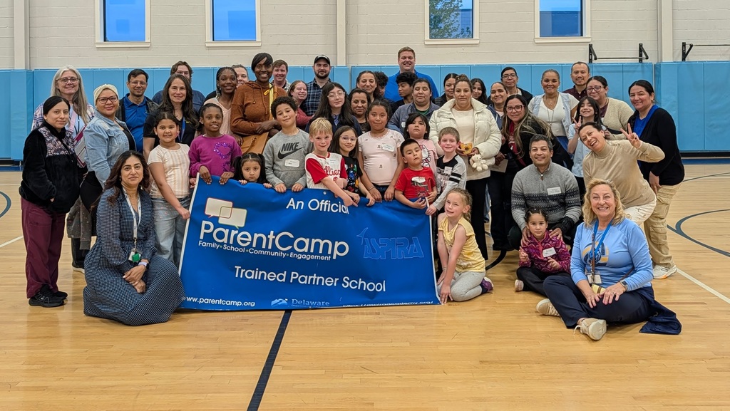 A group of people are posing for a photo in a gym, holding a blue banner that reads "ParentCamp."