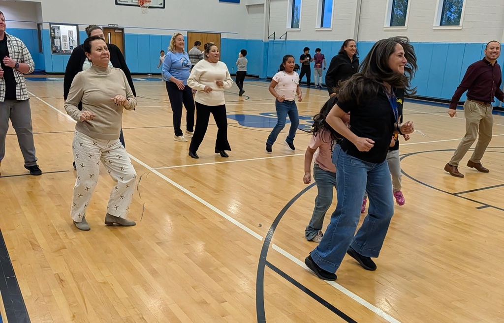 A group of adults and children participate in a dance activity in a gymnasium with a wooden floor.