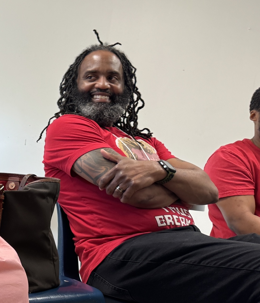 A man with dreadlocks and a beard sits with his arms crossed. He wears a red shirt and has a watch on his left wrist.