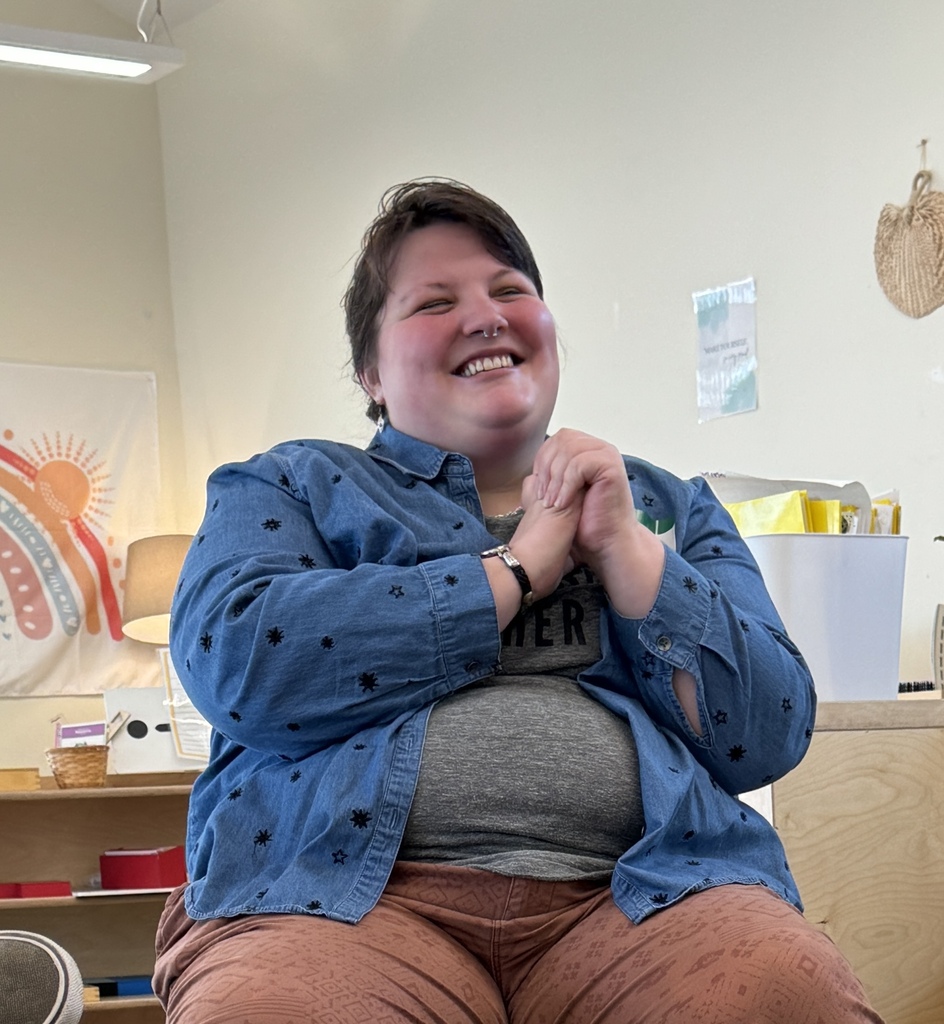 Person in a blue denim jacket and brown pants smiling while sitting in a room.