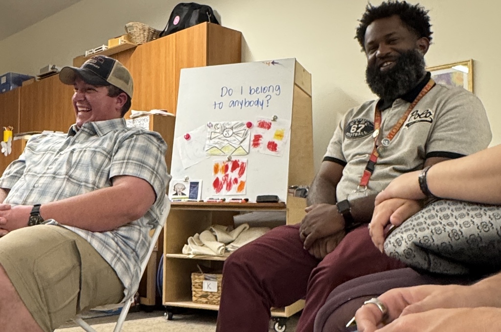 Two men sit next to each other. One wears a cap, and the other has a beard. A whiteboard is behind them.