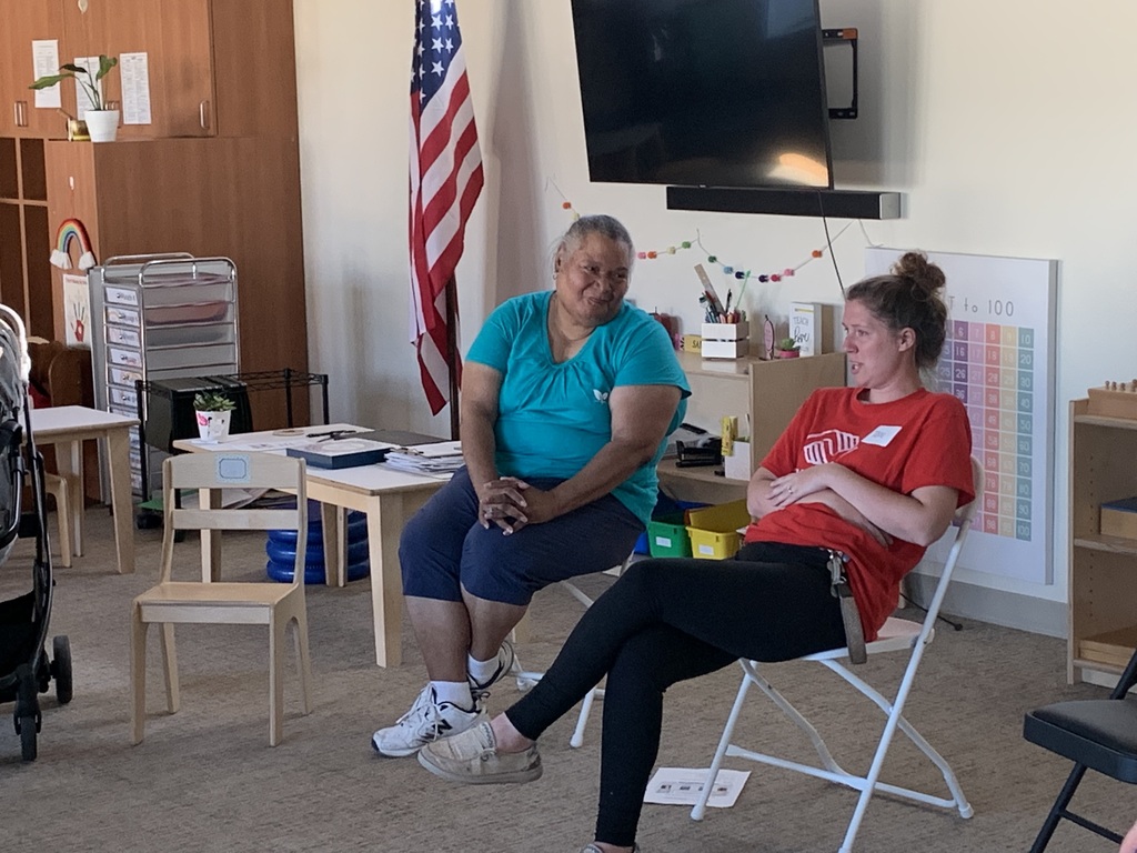 Two women sit on chairs in a room with a table and an American flag. One wears a blue shirt and the other a red one.