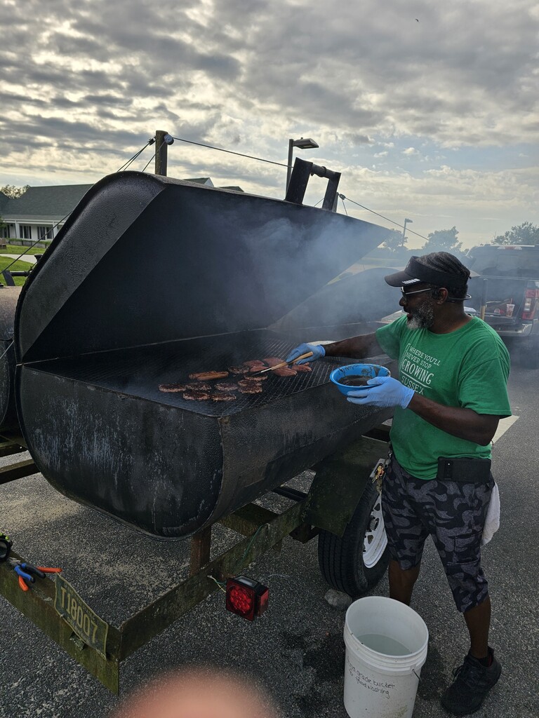 Person in green shirt and blue gloves grilling burgers on a barbecue in an open trailer.