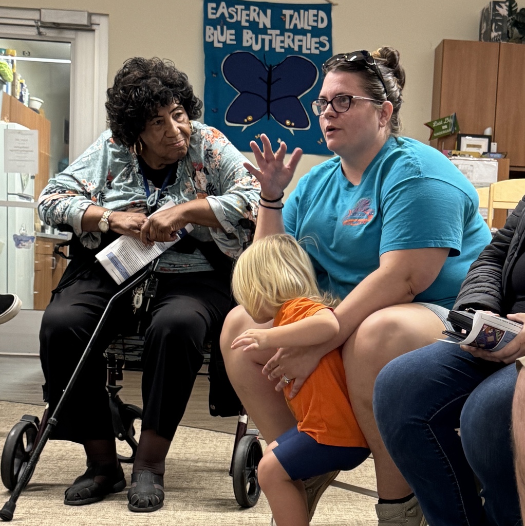 People seated in a room, one woman in a wheelchair speaks to another woman holding a child.