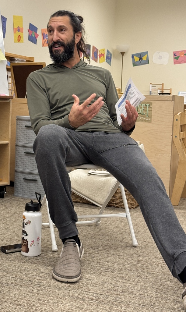A man with a beard sits in a chair, holding a paper, speaking. A water bottle is on the floor.