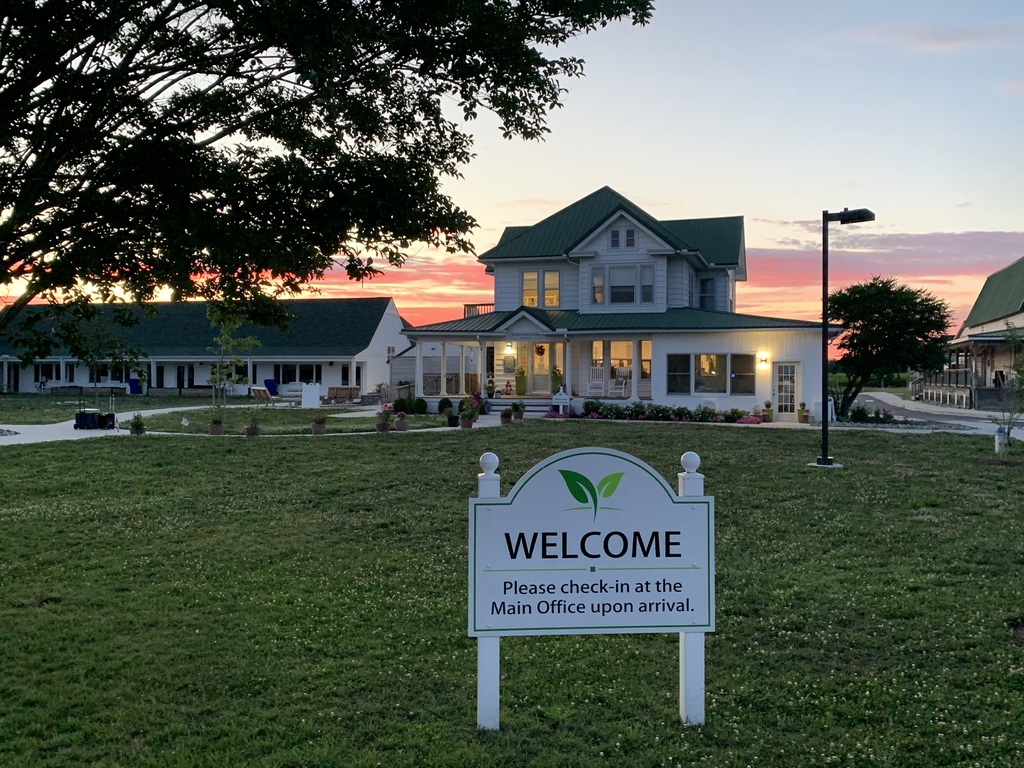 A large house stands with a "Welcome" sign in a grassy field at dusk. Trees and bushes frame the scene.