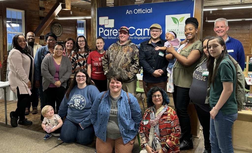 A group of people in a room, some standing, some sitting, and some holding babies, in front of a blue banner.