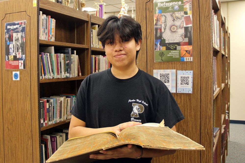 Naasawva holding a very large book in the library, a true scholar