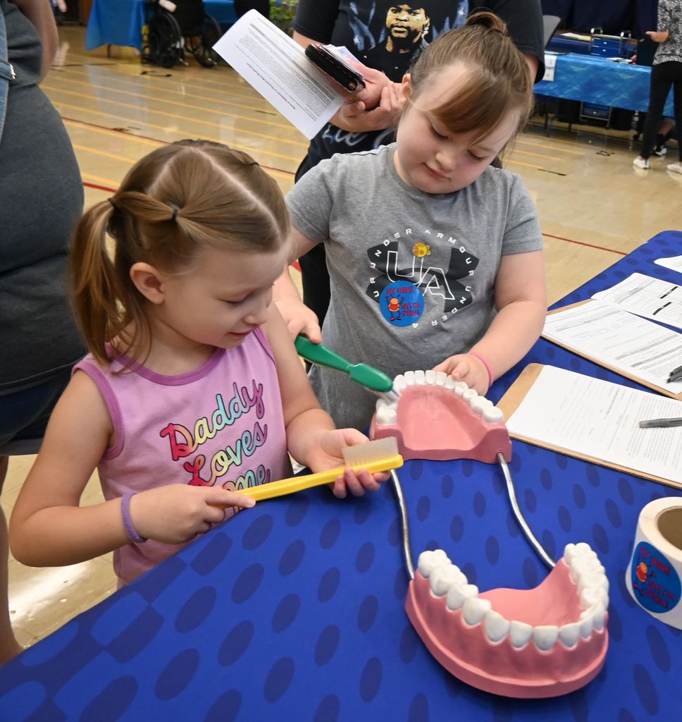 Two sisters stay engaged in brushing the teeth on a model of a mouth at the nurse's station.
