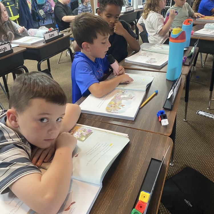A student reading from a textbook using a microphone while other students listen