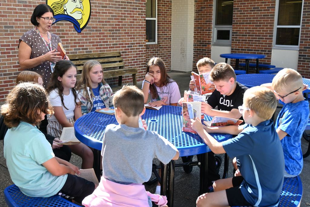 Students sit outside at a picnic table to read with Mrs. Amrstrong.