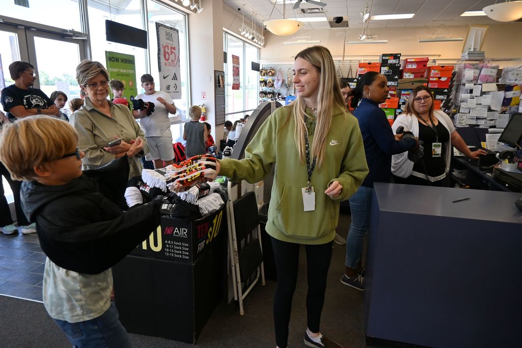 A boy is handed a package of socks and boxes of shoes for kids loom in the background.
