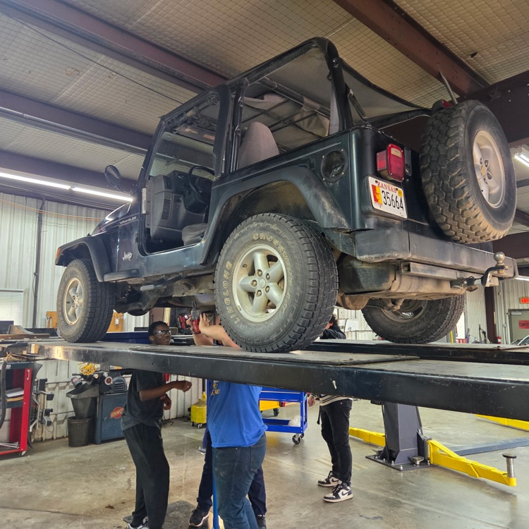 Students work under a jeep raised up on a lift.