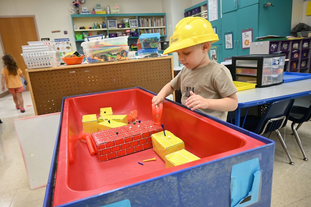 A little boy wears a plastic hard hat and hammers golf tees into cardboard rectangles.