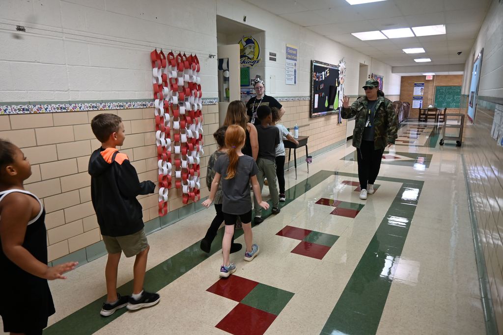 A teacher wearing camo and acting as mild mannered  drill sergeant leads a group of students into her room.