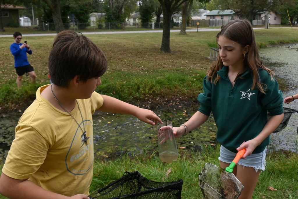 A boy puts a little minnow in a 2 liter container a girl is holding.