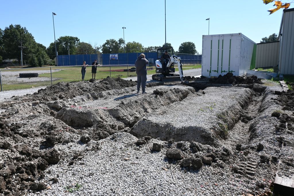 As students look on, Mr. Gilpin operates a mini excavator to dig out the footings for the greenhouse.