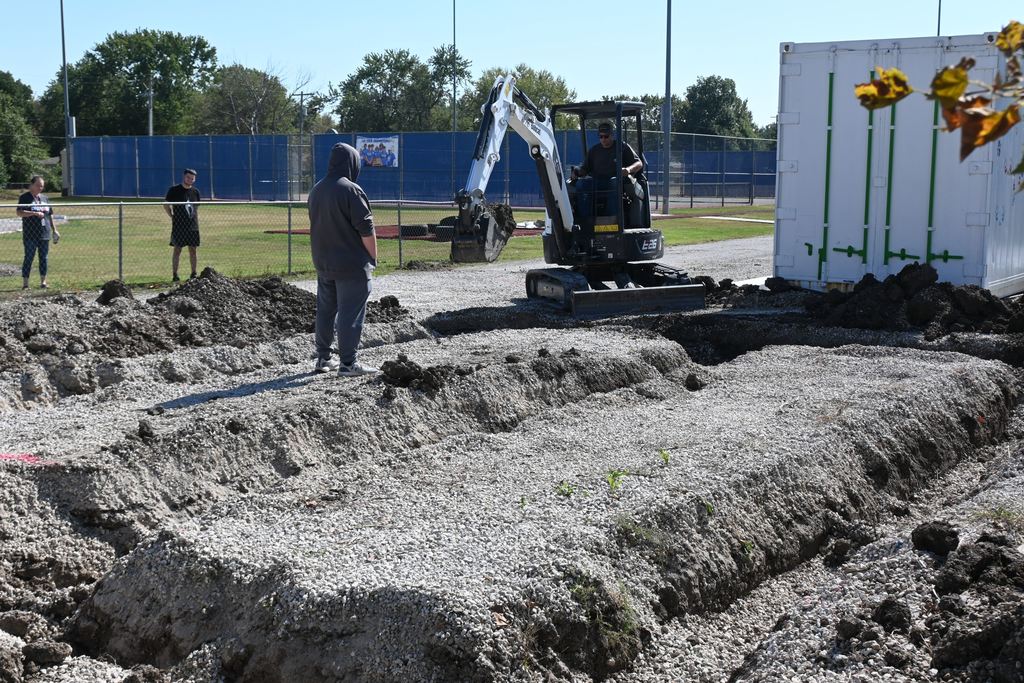 As students look on, Mr. Gilpin operates a mini excavator to dig out the footings for the greenhouse.